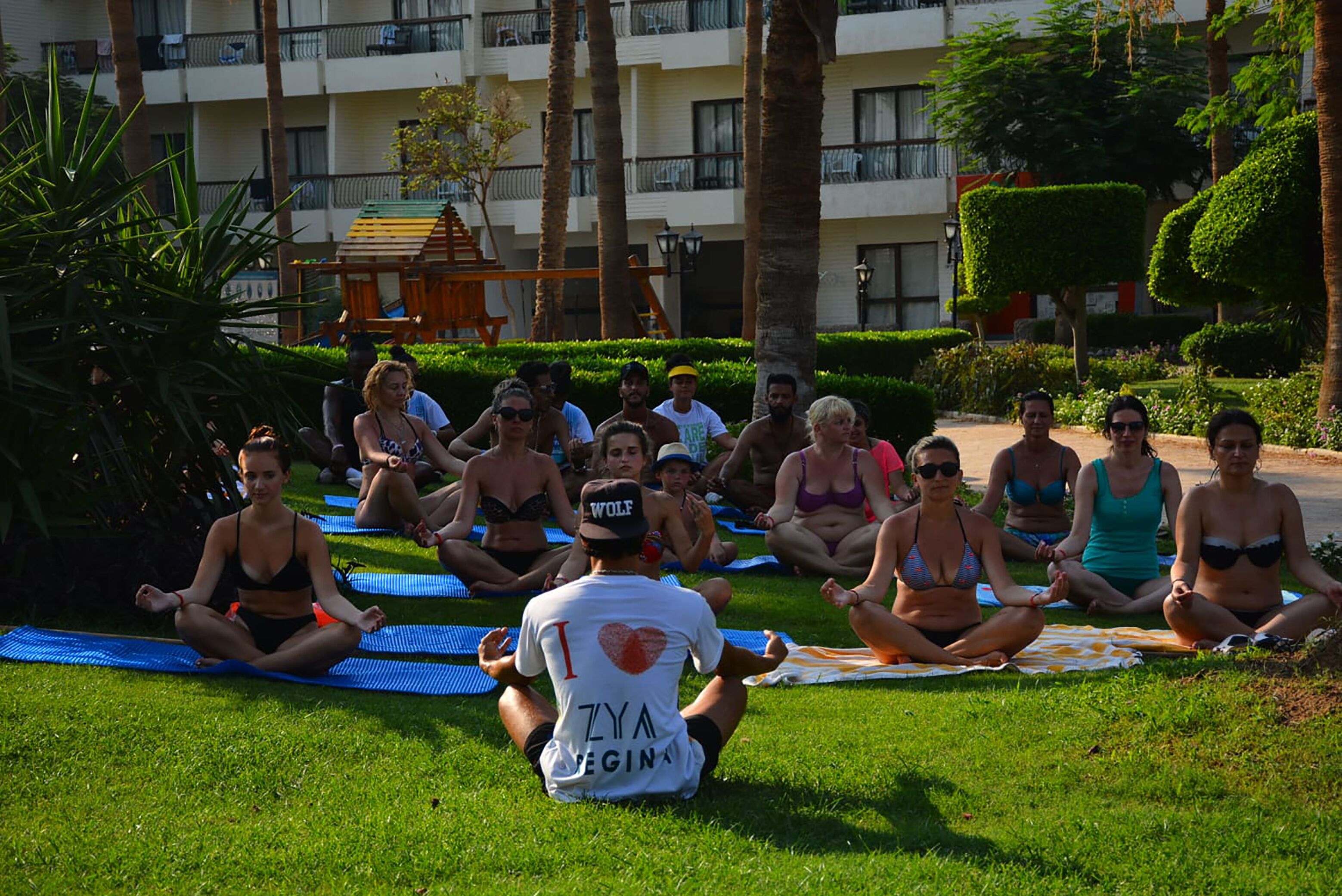 A group of people sitting on yoga mats outdoors in a grassy area with a building in the background.