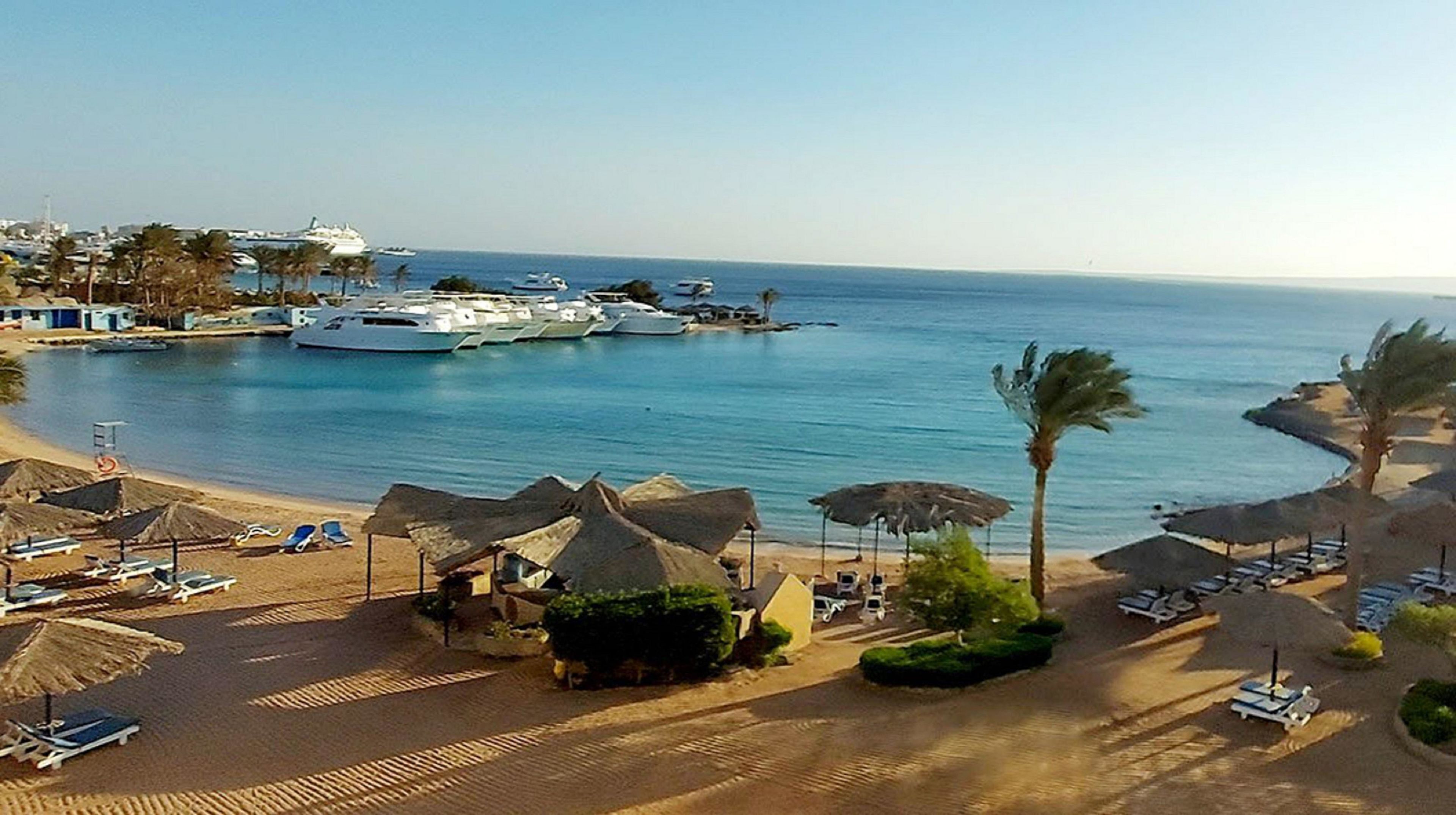 Una playa con tumbonas, sombrillas y un puerto deportivo con barcos.