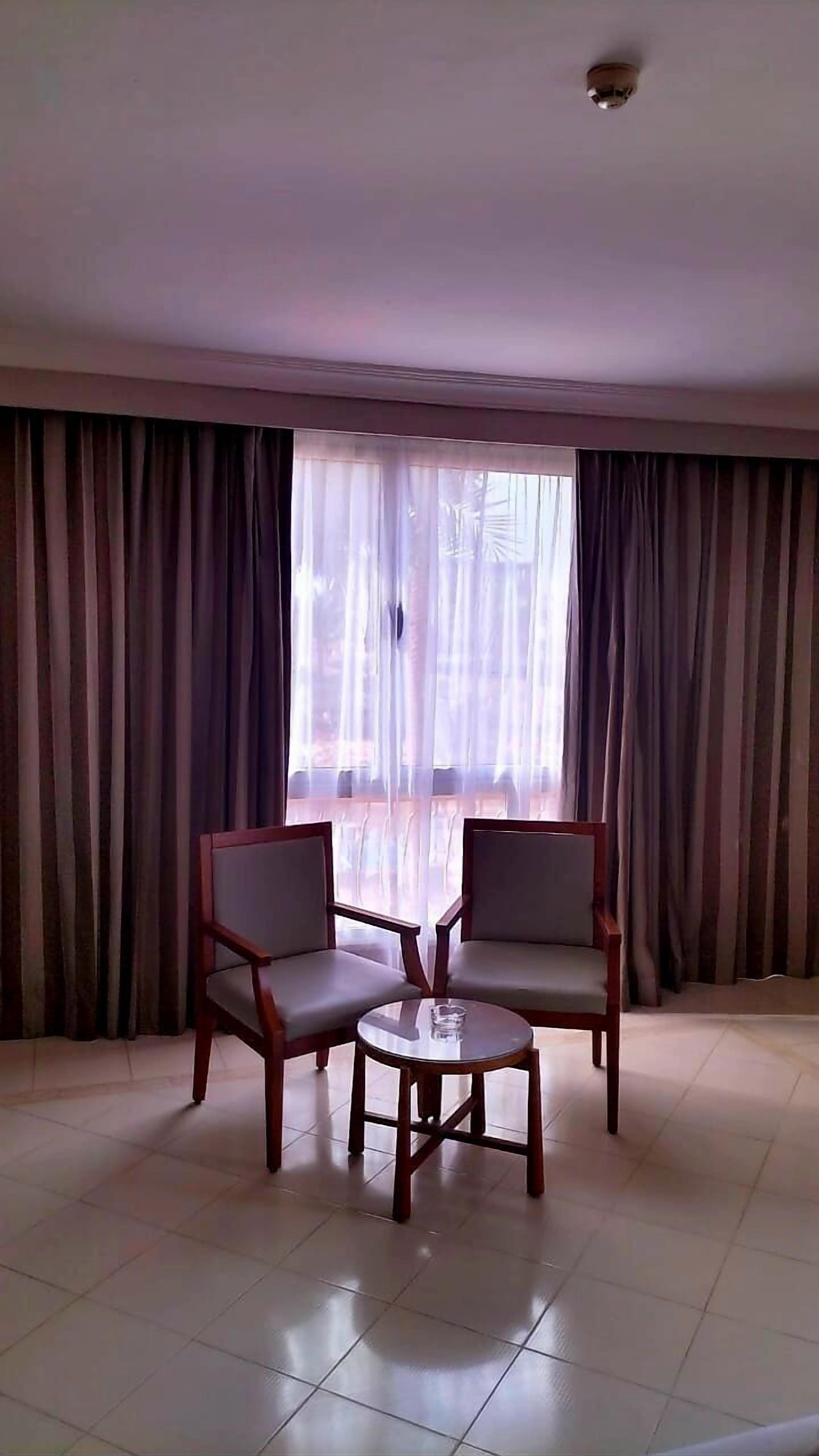 A room with two wooden chairs and a small round table, large windows with sheer curtains, and a ceiling light.
