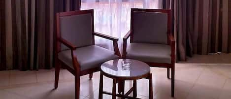 A room with two wooden chairs and a small round table, large windows with sheer curtains, and a ceiling light.