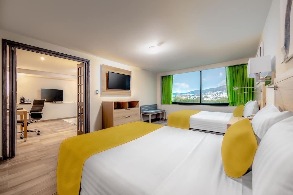 Une chambre d’hôtel équipée d’un lit, d’un bureau, d’une chaise, d’une télévision et d’une fenêtre donnant sur un paysage urbain.