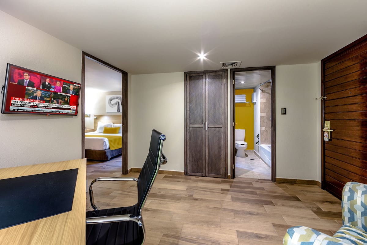 Une chambre d’hôtel avec un plancher en bois, une chaise noire, un canapé à motifs, une porte en bois et un téléviseur diffusant un programme d’information.