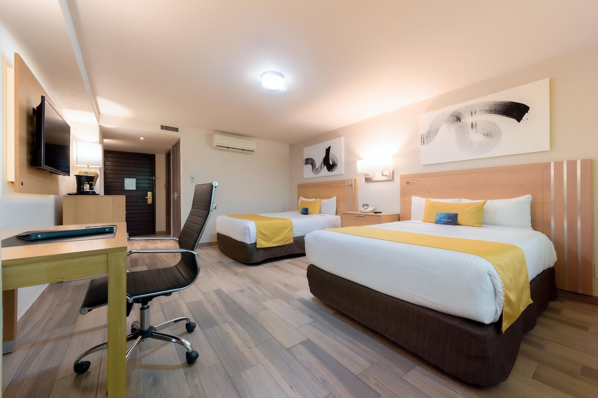 A hotel room with two beds, a desk, a chair, a TV, and artwork on the walls.