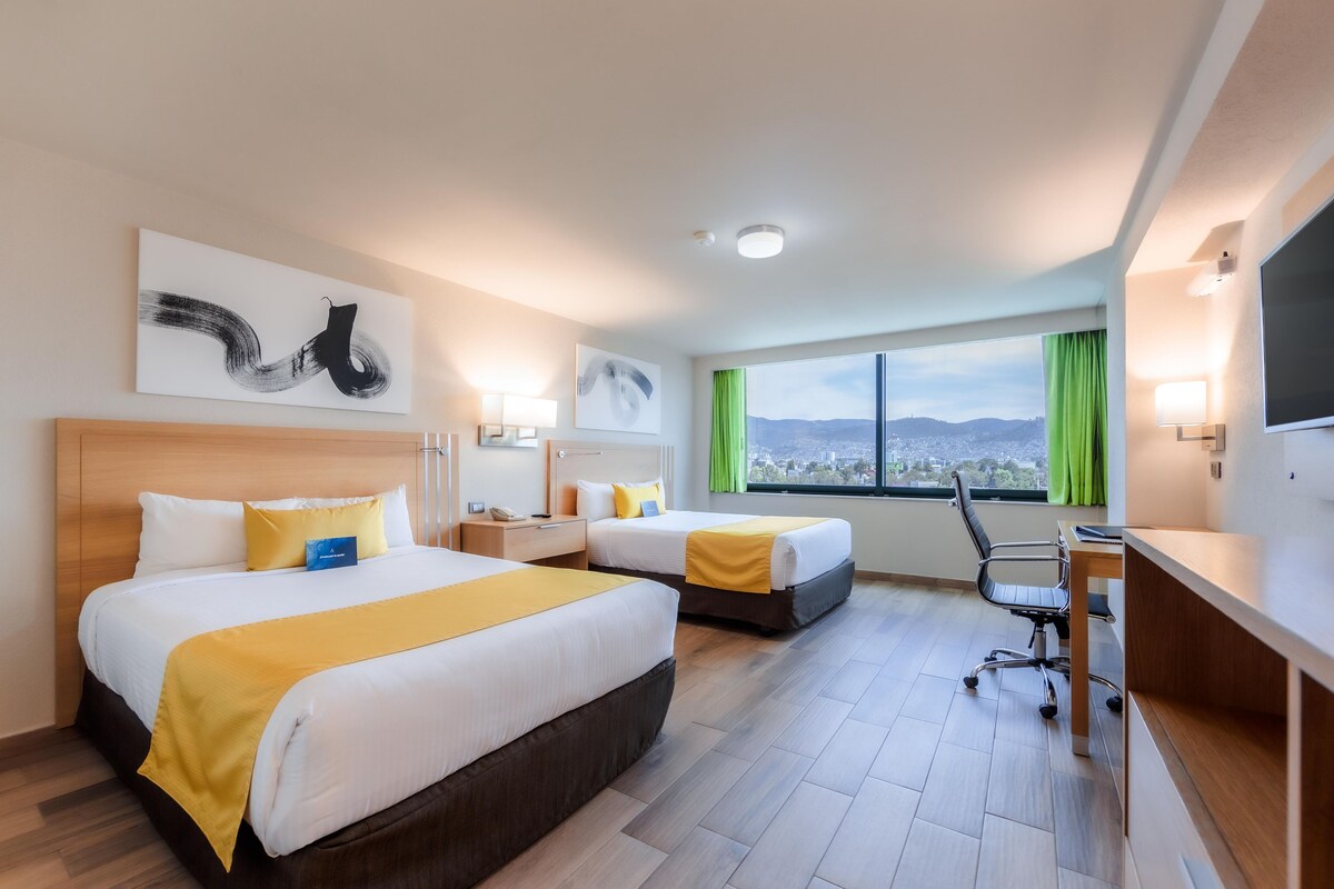 A hotel room with two beds, a desk, a chair, and a large window with a city view.