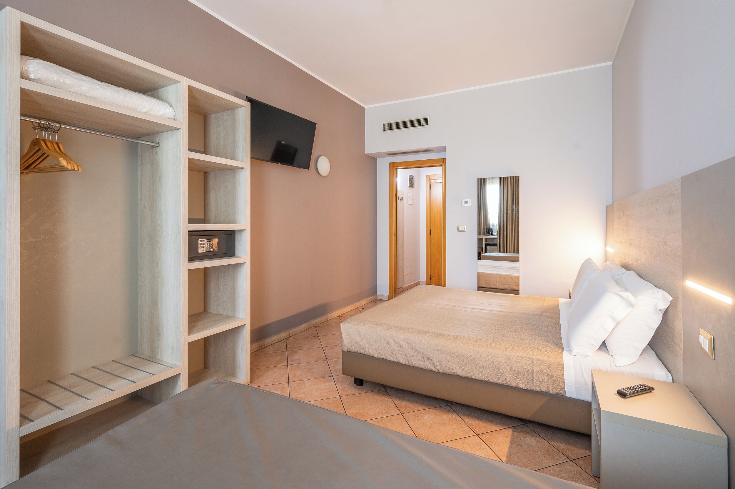 Un dormitorio con una cama, una mesita de noche, un televisor montado en la pared, un armario y una puerta que da a otra habitación.