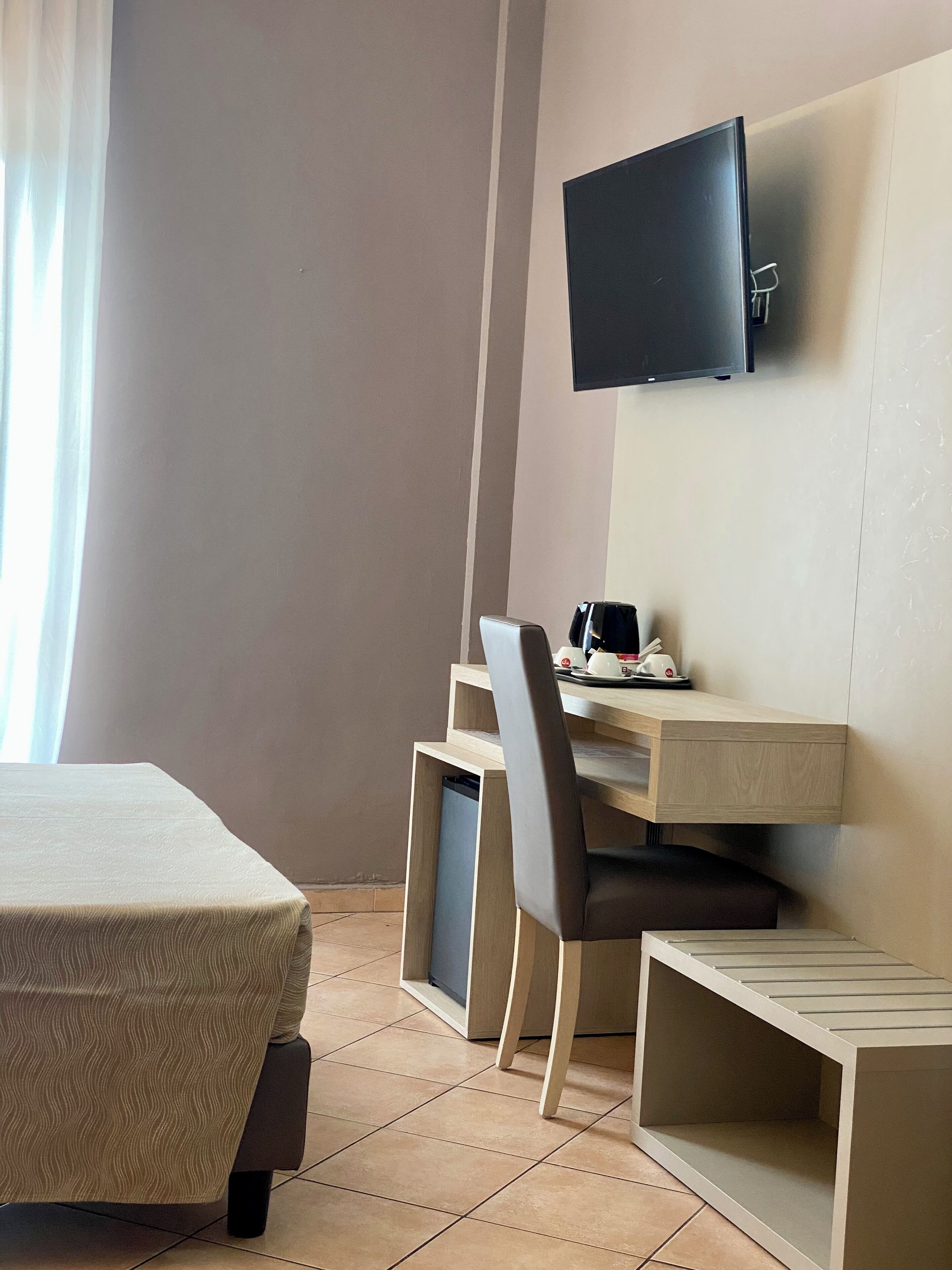A hotel room with a bed, a desk with a chair, a television mounted on the wall, and a window with curtains.