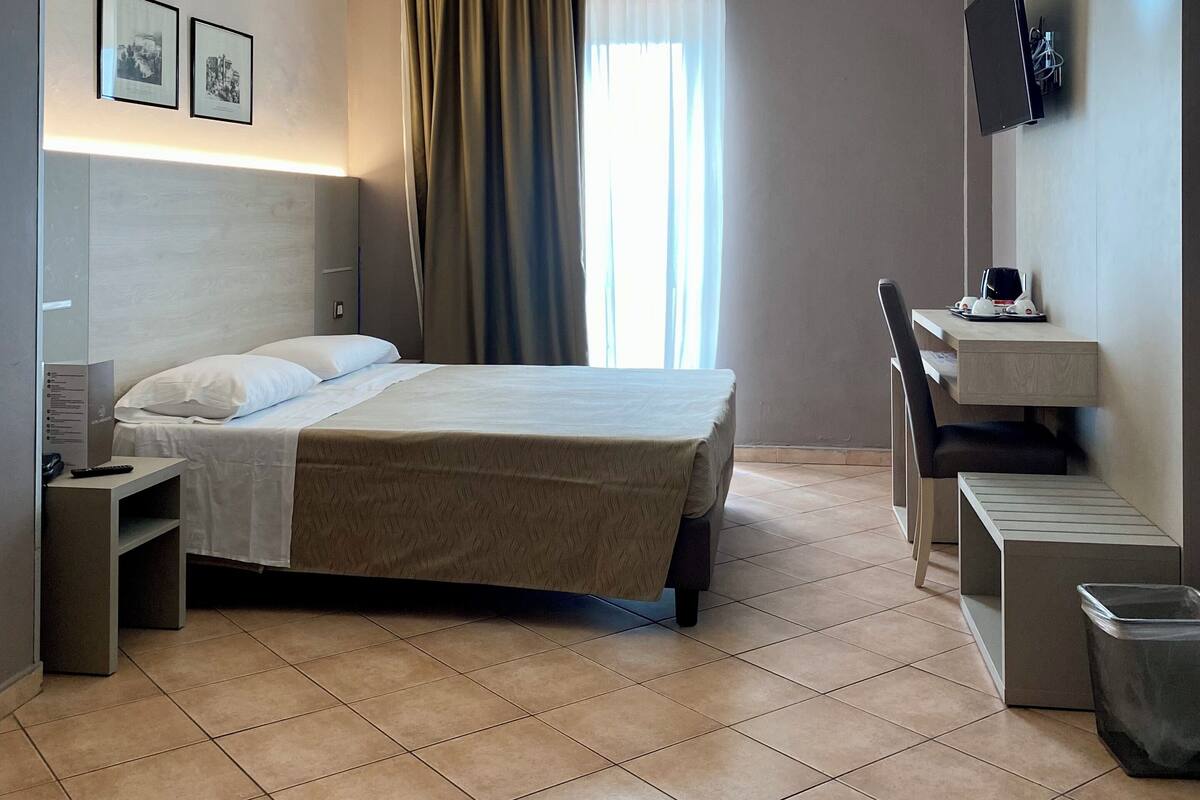 A hotel room with a bed, bedside table, TV, desk, chair, and a trash bin.