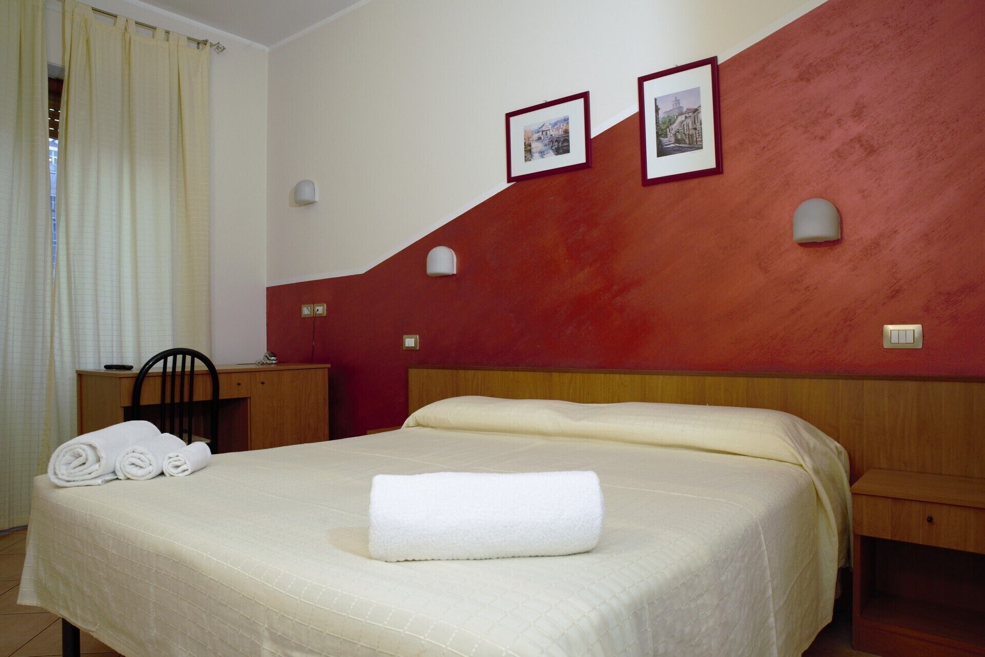 A hotel room with a large bed, a wooden nightstand, a desk, and two wall-mounted lamps.