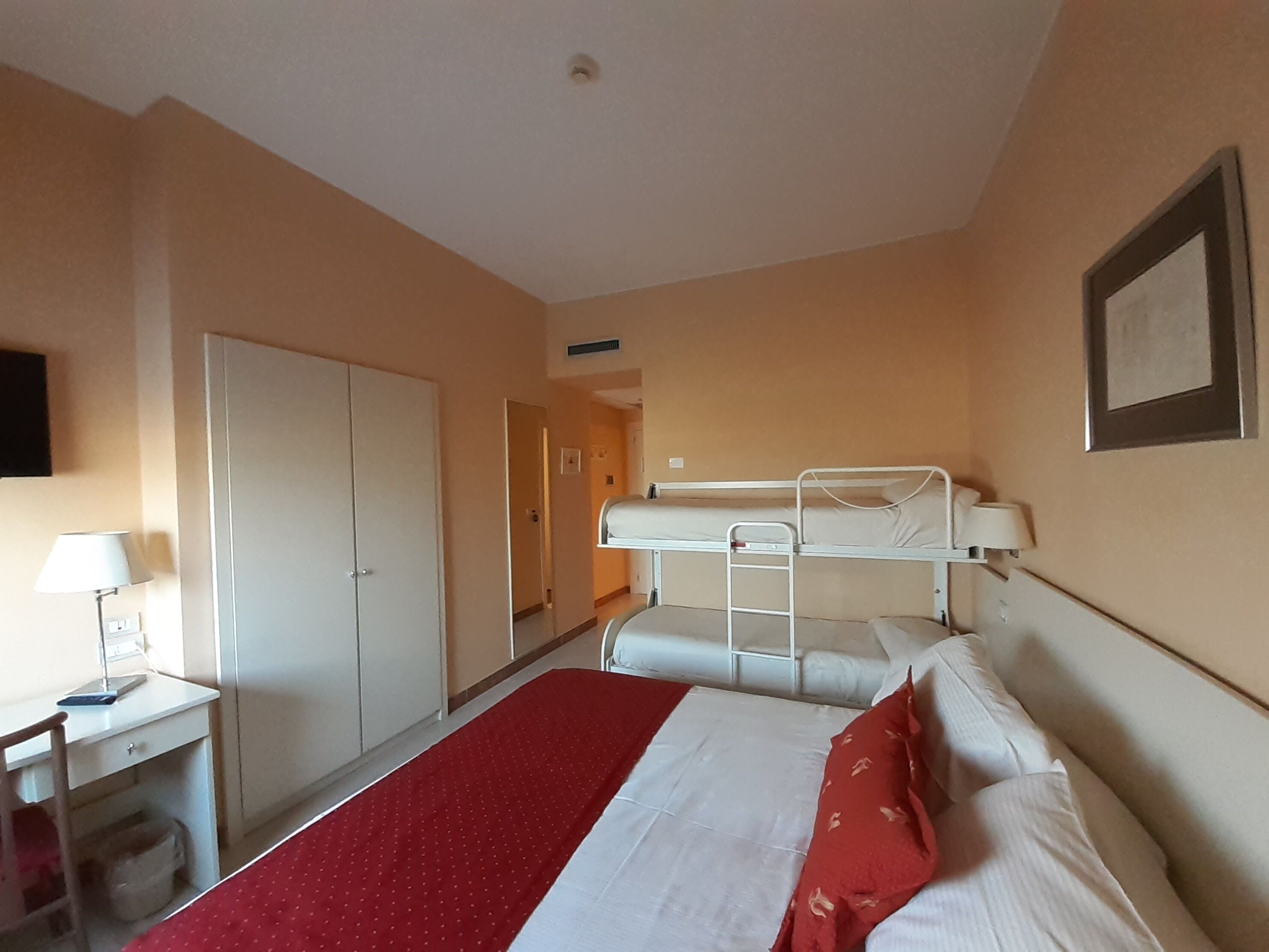 A hotel room with a bunk bed, a desk with a lamp, a wardrobe, and a door leading to another room.