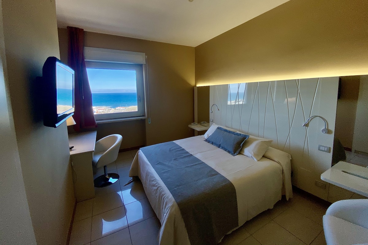 Habitación de hotel con cama, escritorio con televisor, silla, mesita de noche y vista al mar.