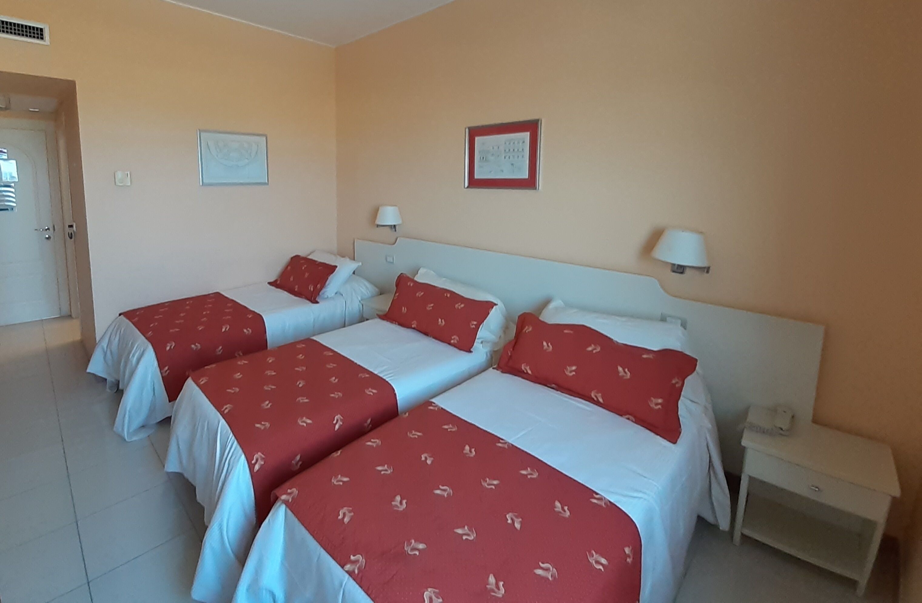 A hotel room with three beds, each with red and white bedding. There are two bedside tables with lamps, a door, and two framed pictures on the wall.