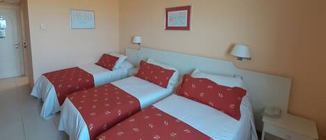 A hotel room with three beds, each with red and white bedding. There are two bedside tables with lamps, a door, and two framed pictures on the wall.