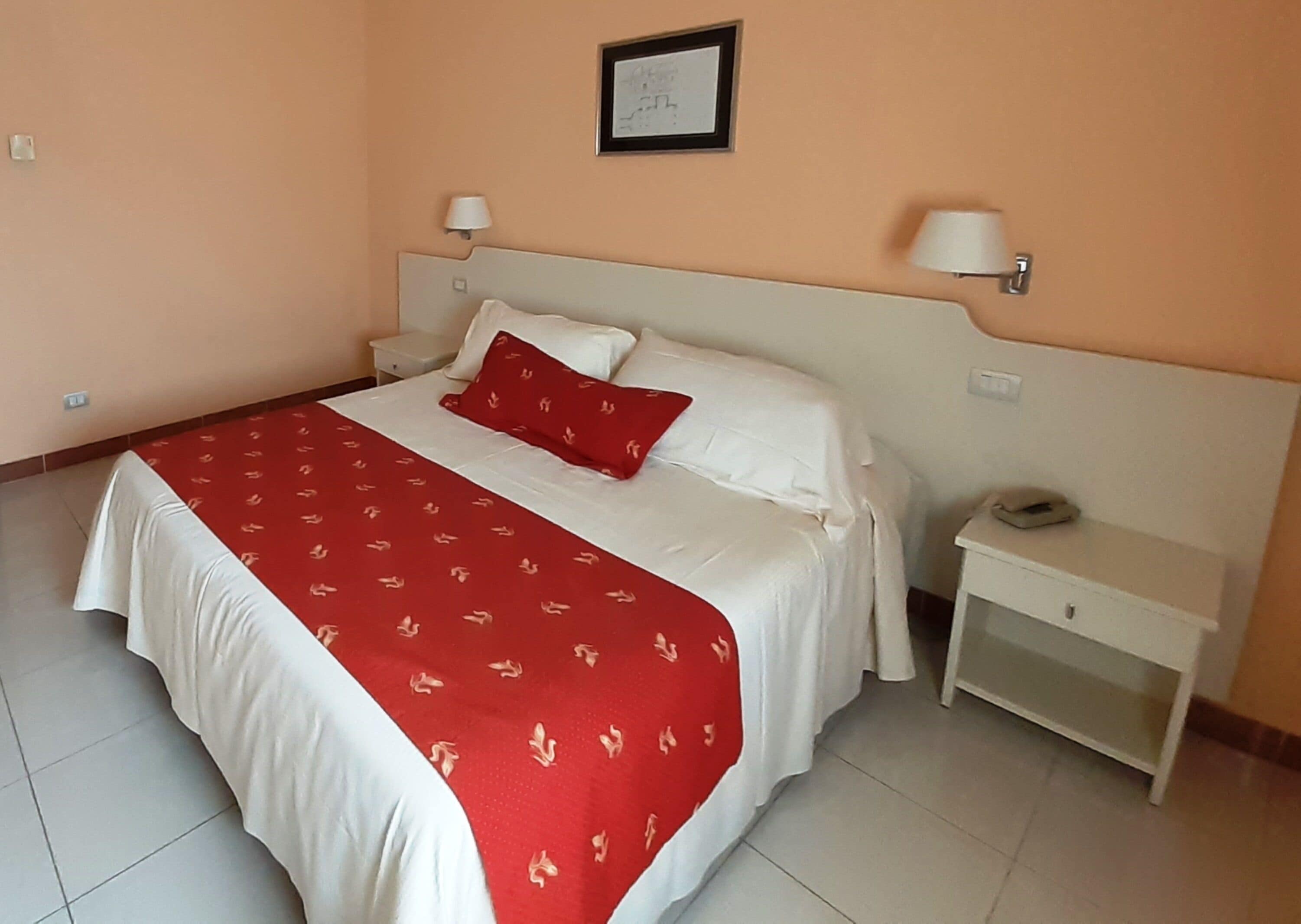 Una cama bien tendida con una colcha blanca y roja, una almohada roja y una mesita de noche blanca con una lámpara.