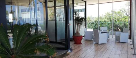 Un espace intérieur moderne doté d’un ascenseur en verre, de plantes en pot et d’un coin salon extérieur avec des chaises blanches.