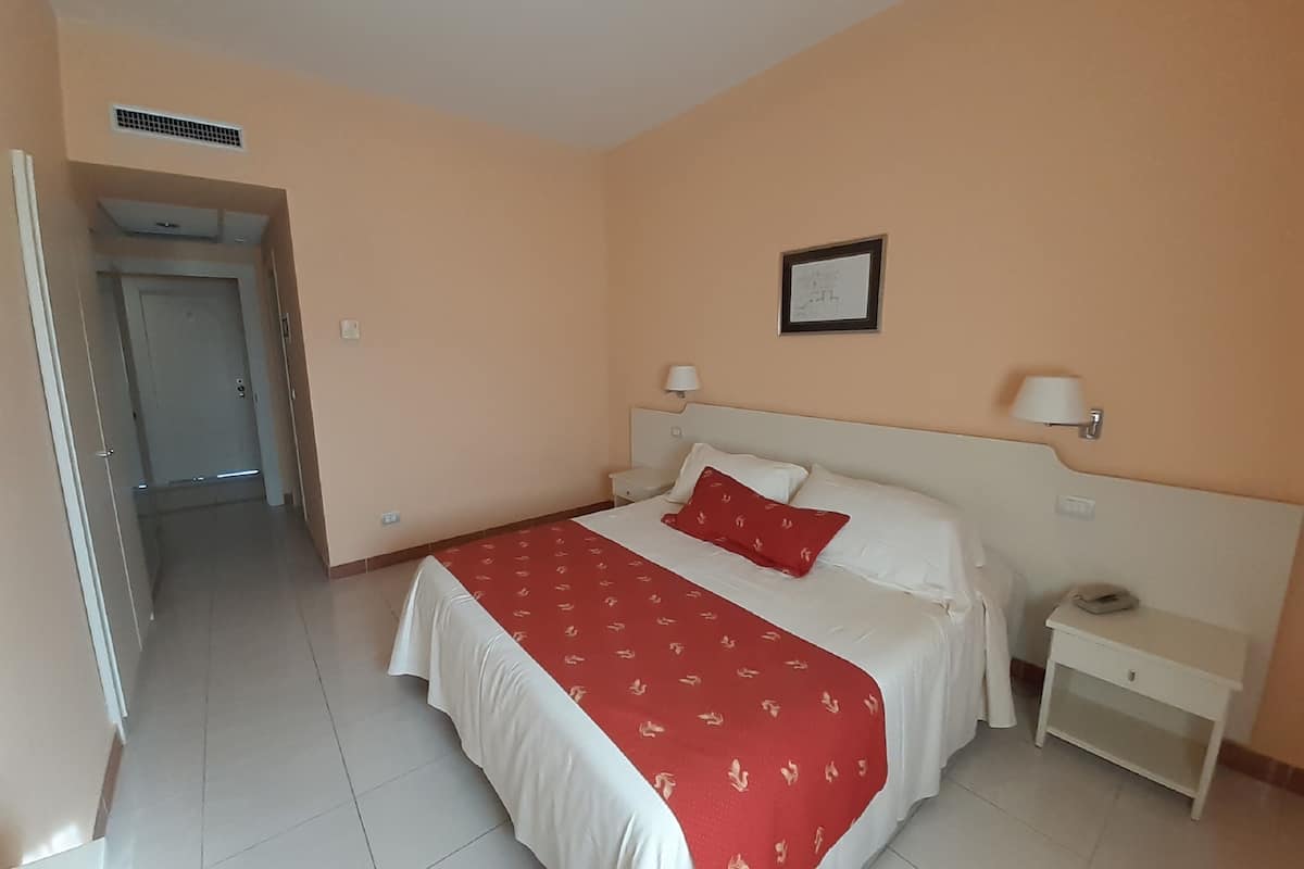 Habitación de hotel con cama, mesitas de noche, lámpara de pared y puerta que da a otra habitación.