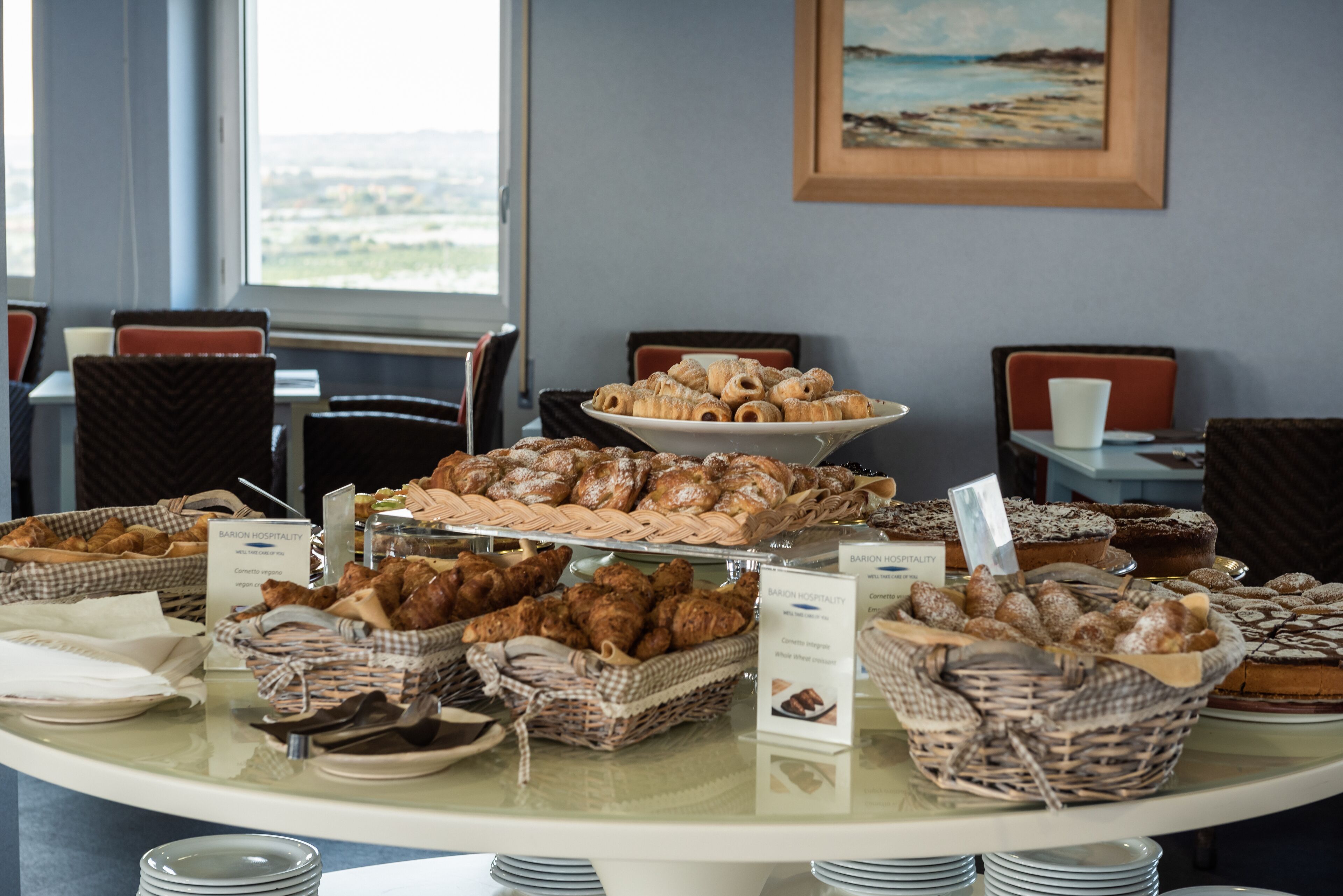 Una mesa de buffet con diversos productos de panadería y pastelería, incluyendo croissants y magdalenas, dispuesta sobre una mesa de cristal en un comedor con sillas y un cuadro en la pared.