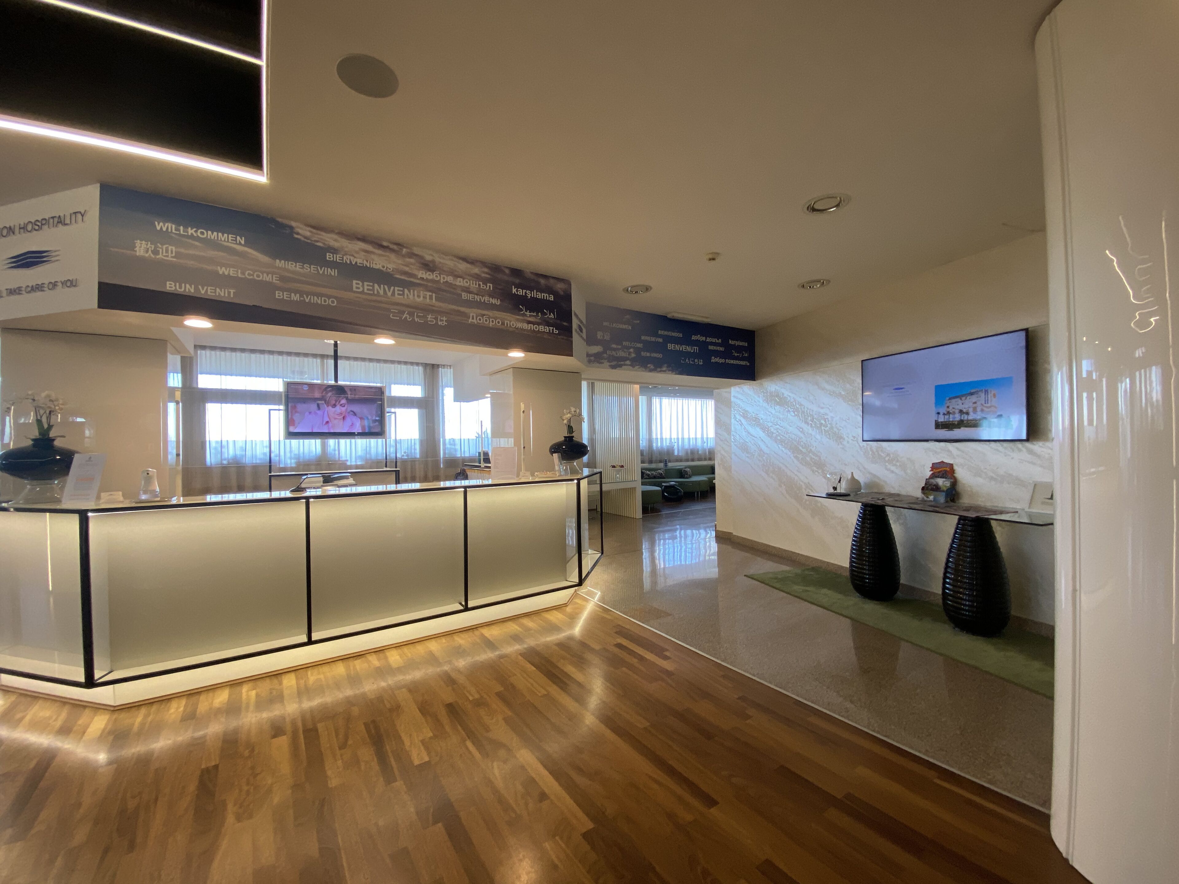 A reception area with a modern design, featuring a reception desk, a television screen, and a large welcome sign with multilingual greetings.