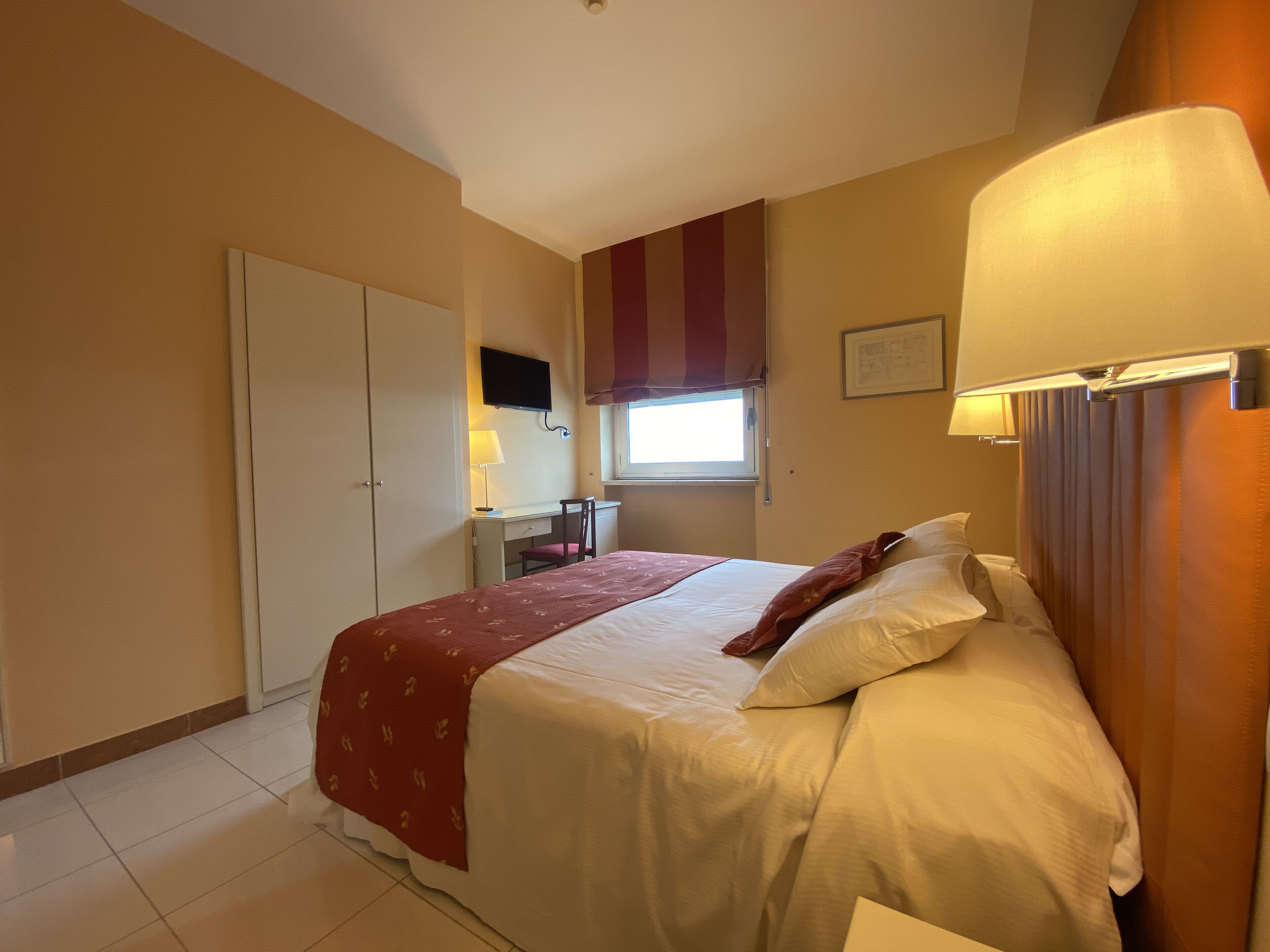 Een slaapkamer met een bed, een bureau, een stoel, een televisie en een raam met gordijnen.