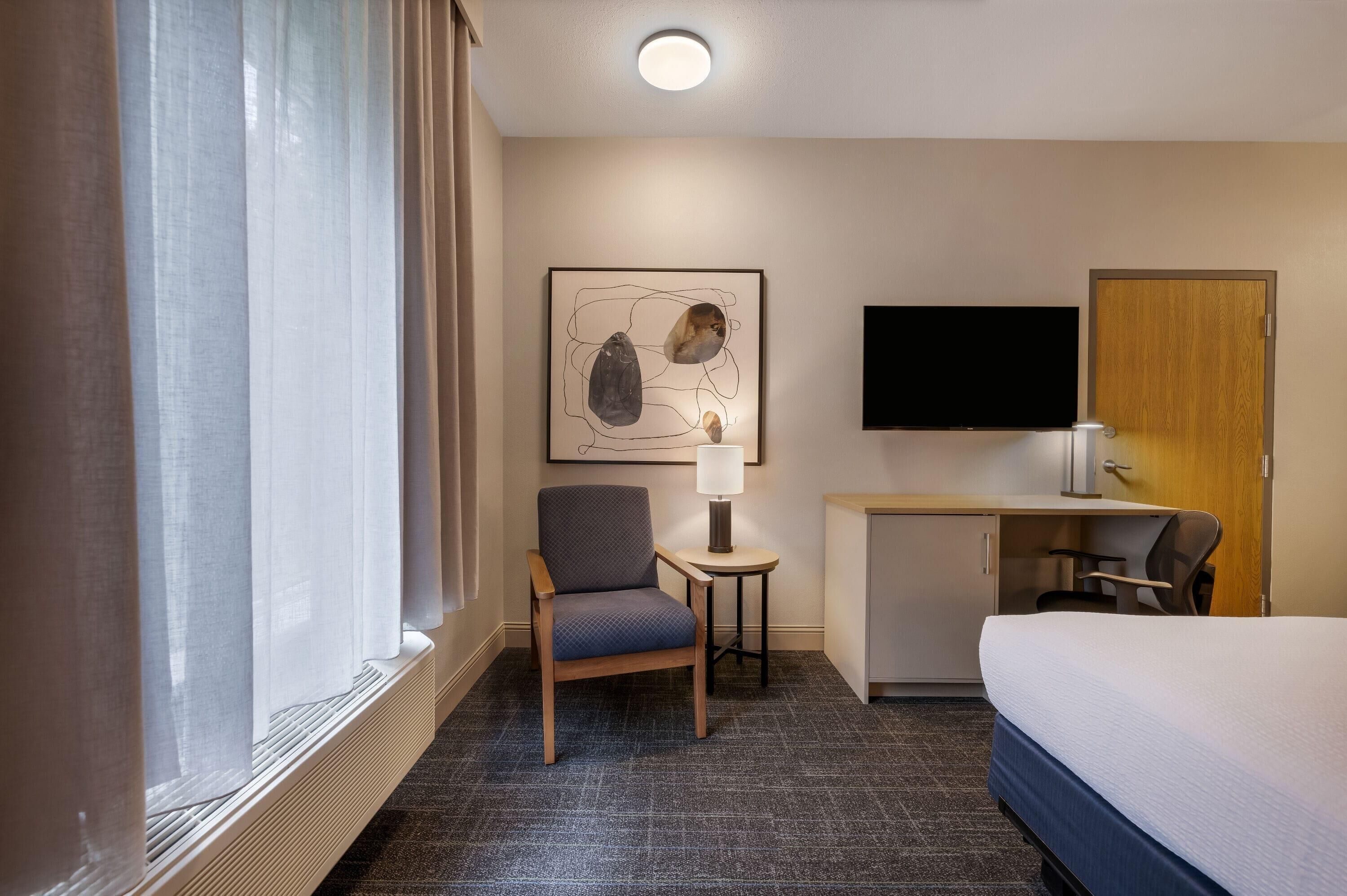 A hotel room with a bed, a desk with a chair, a TV, and a painting on the wall.