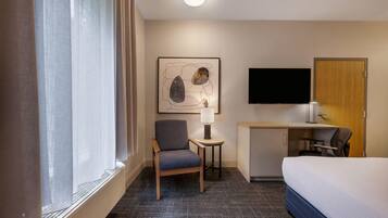 A hotel room with a bed, a desk with a chair, a TV, and a painting on the wall.