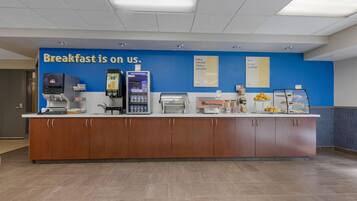 A breakfast station with a blue wall, a coffee machine, a milk dispenser, and a sign that reads 'Breakfast is on us.'