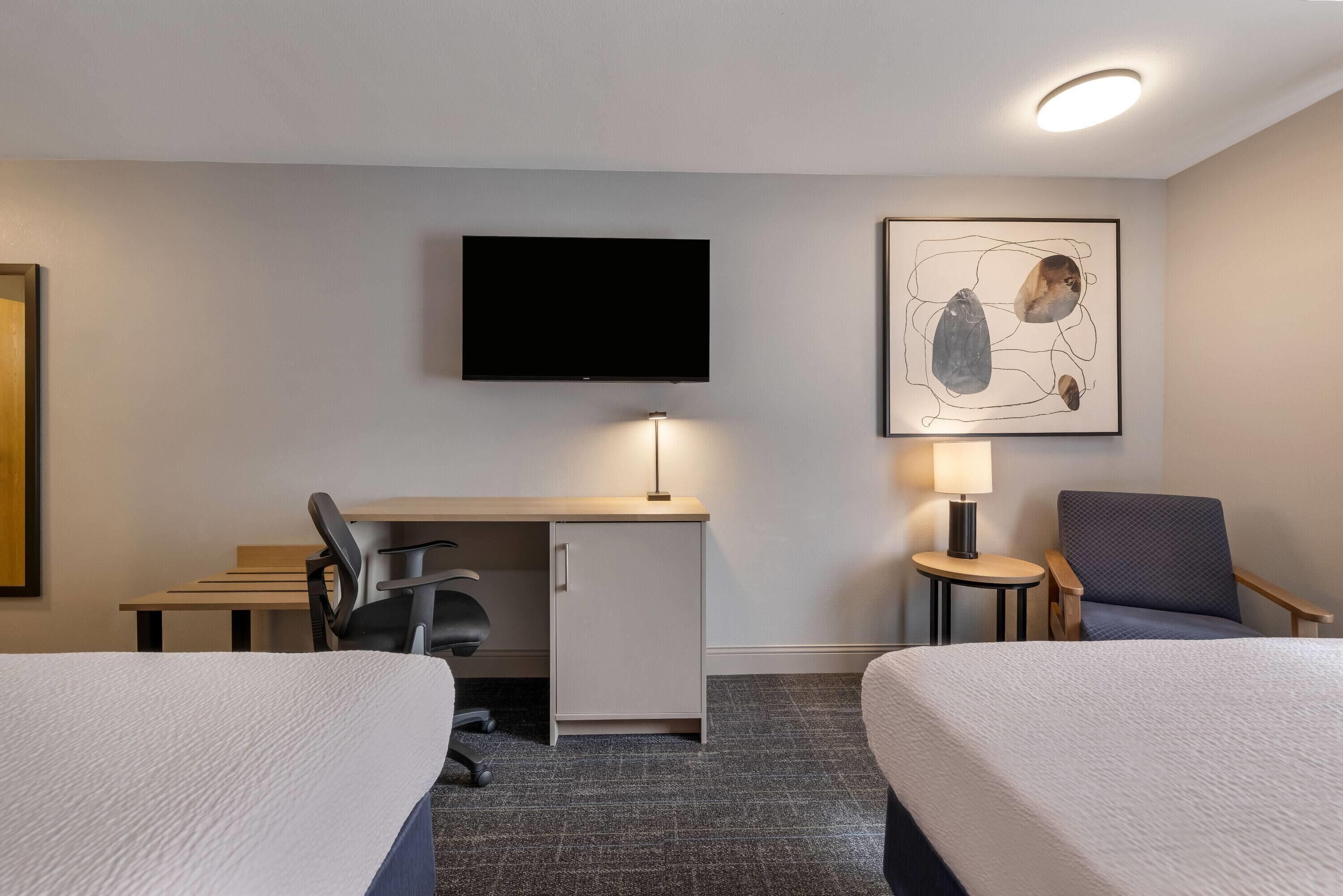A hotel room with two beds, a desk, a chair, a television, and a lamp.