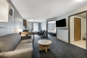 Habitación de hotel con sofá, una mesita, televisor montado en la pared y puerta que da al baño.