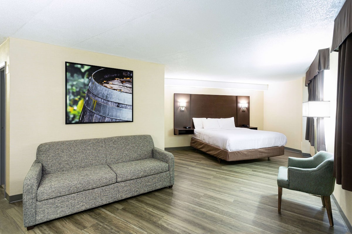 A hotel room with a sofa, a bed, a chair, a nightstand, a lamp, and a painting on the wall.