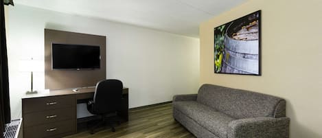 Una habitación de hotel con un escritorio de madera, una silla de oficina negra, un sofá gris, un televisor de pantalla plana montado en la pared y un cuadro con una barrica.