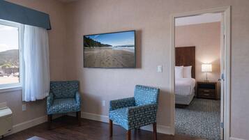 Habitación de hotel con un ventanal grande, una cama, una mesita de noche, una silla y un televisor de pared mostrando una escena de playa.