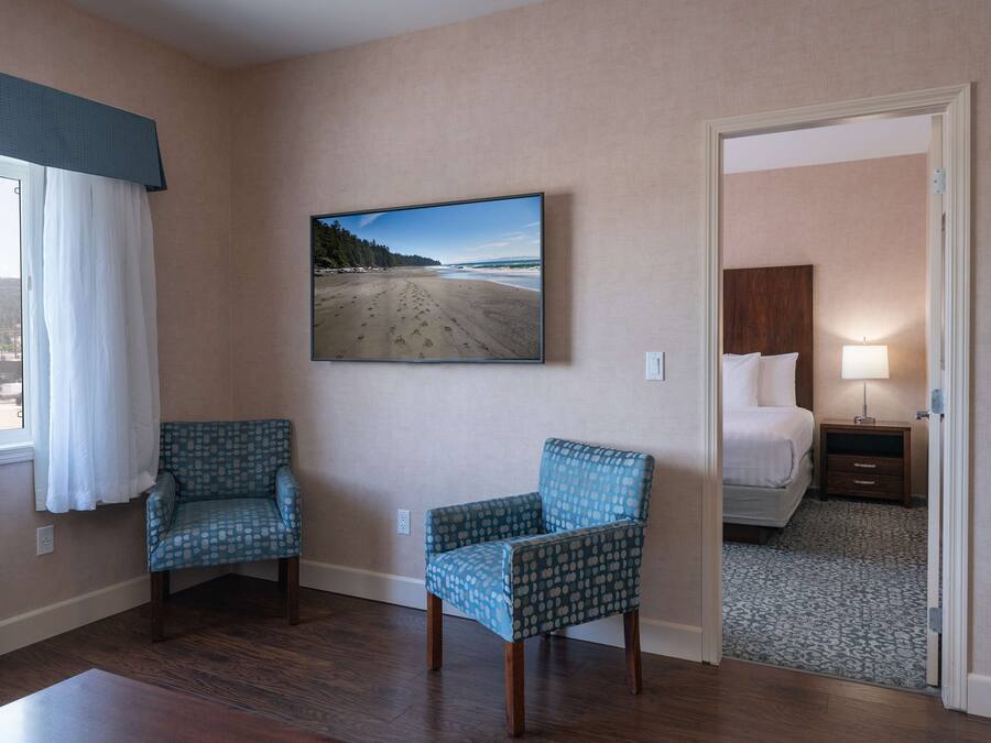 Habitación de hotel con un ventanal grande, una cama, una mesita de noche, una silla y un televisor de pared mostrando una escena de playa.