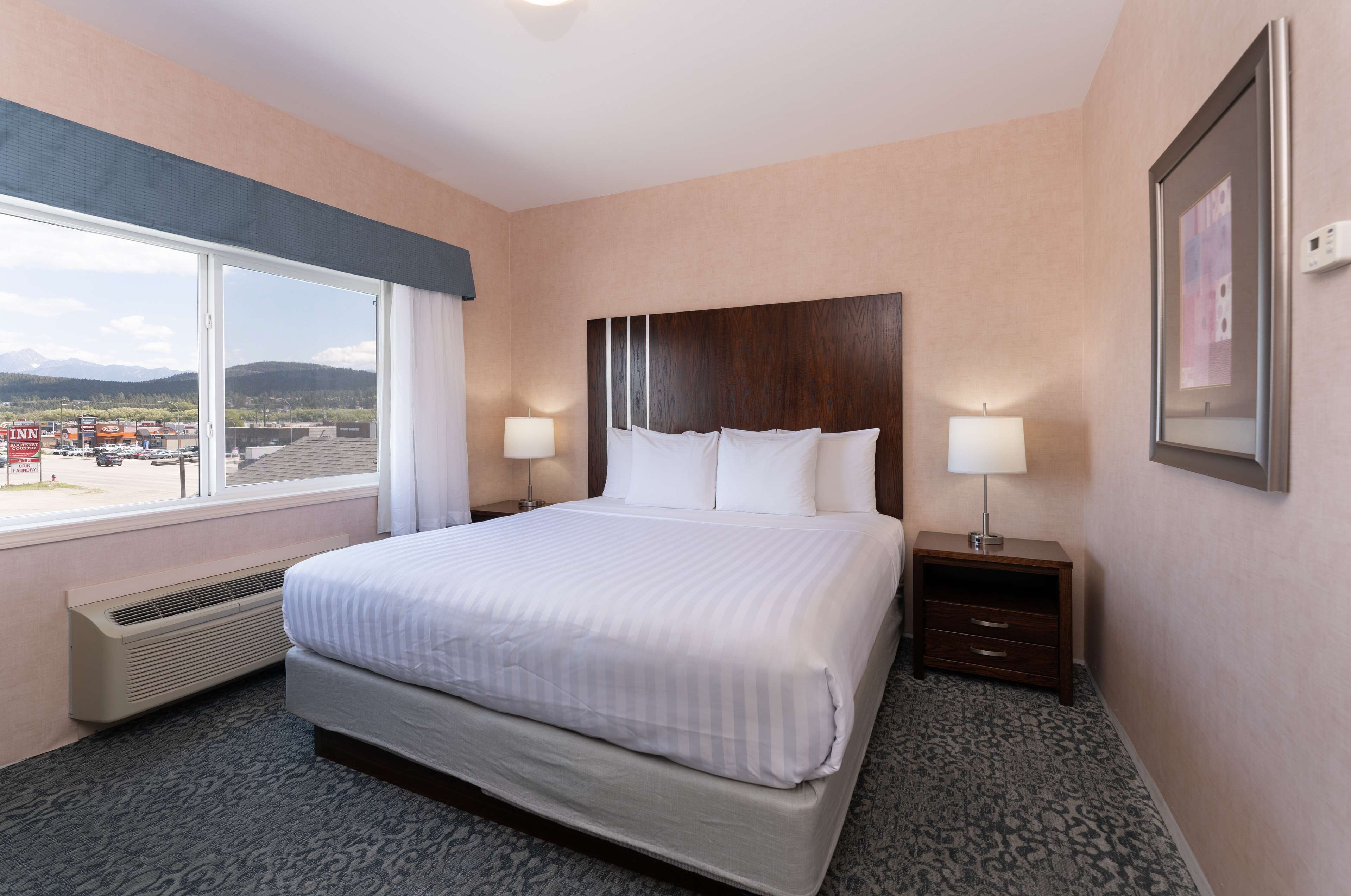 A hotel room with a large bed, two bedside tables with lamps, a window with a view of mountains, and a framed picture on the wall.