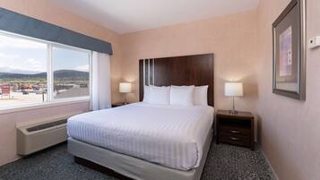 A hotel room with a large bed, two bedside tables with lamps, a window with a view of mountains, and a framed picture on the wall.