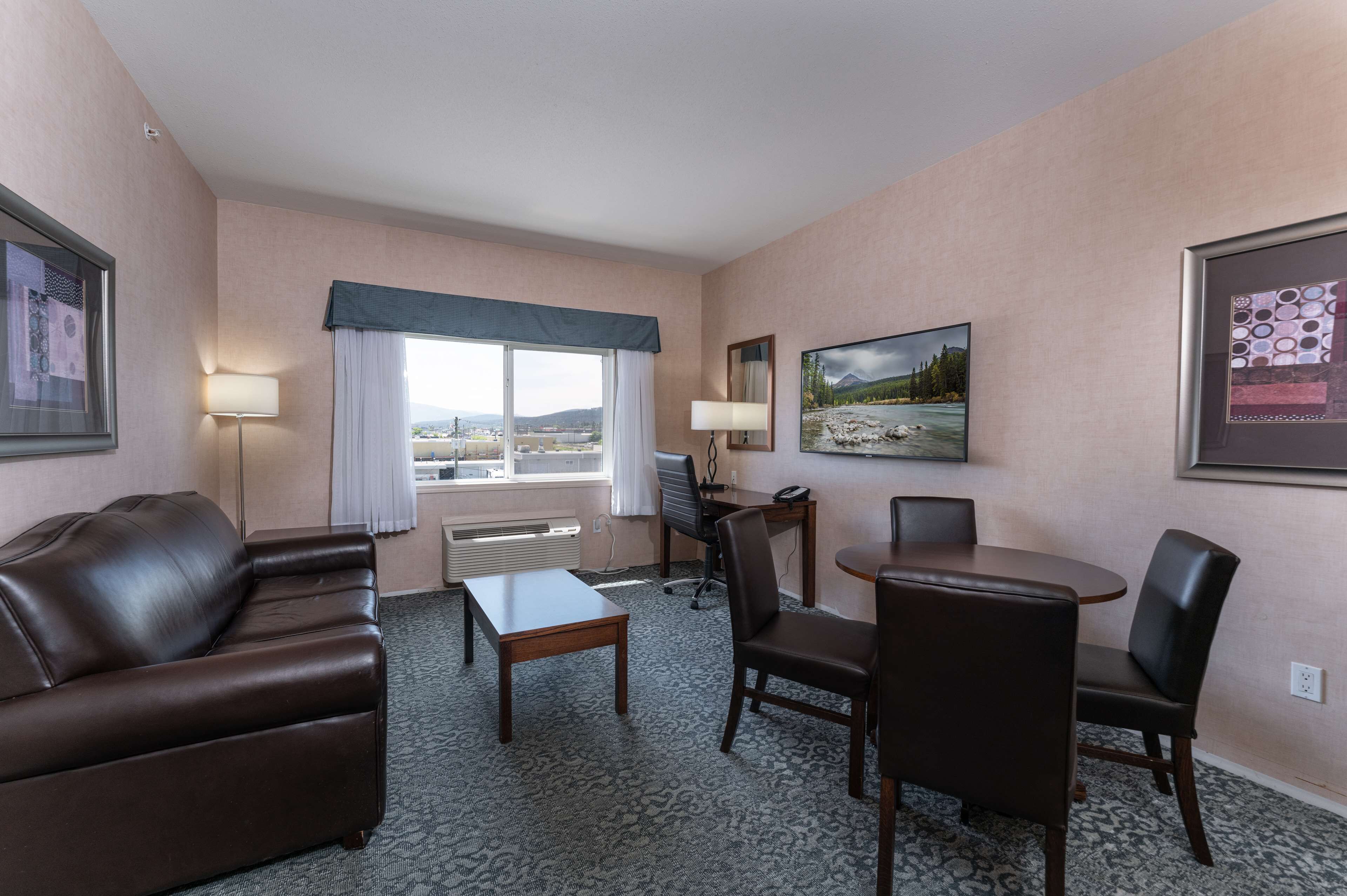 A hotel room with a leather sofa, a small table, a dining table with chairs, a desk with a chair, a large window with a view of the outdoors, and two framed pictures on the walls.