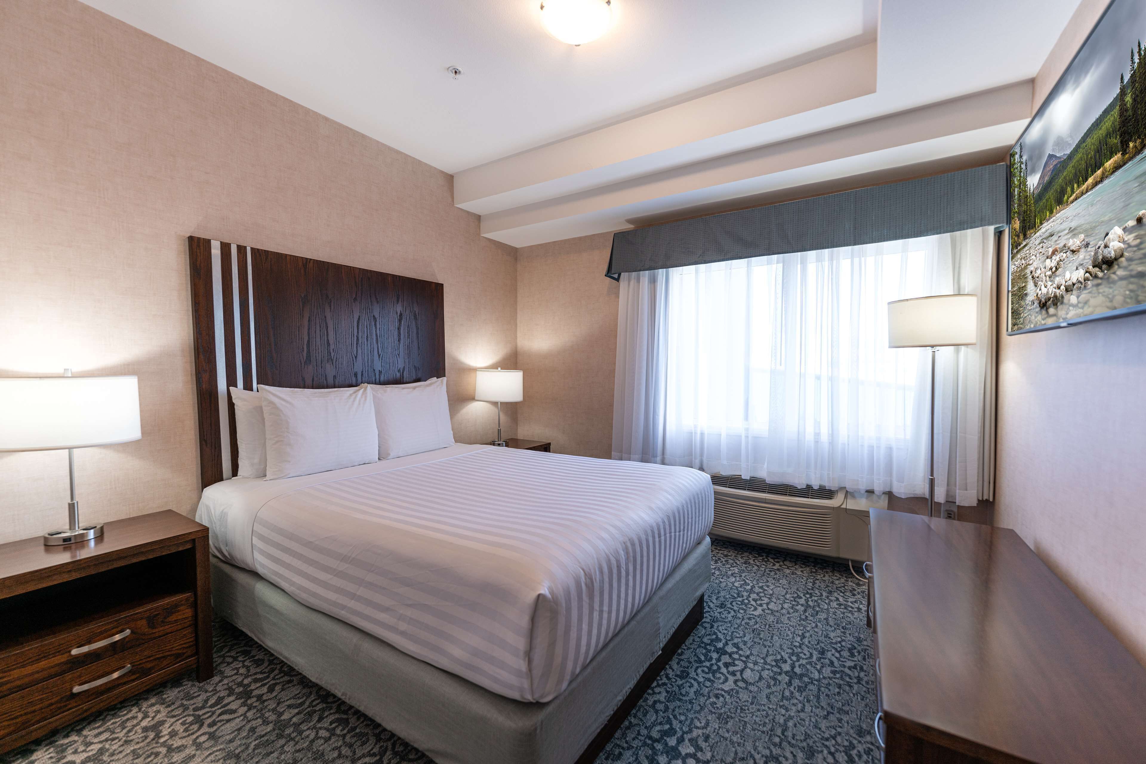A hotel room with a large bed, two bedside tables with lamps, a window with curtains, and a painting on the wall.