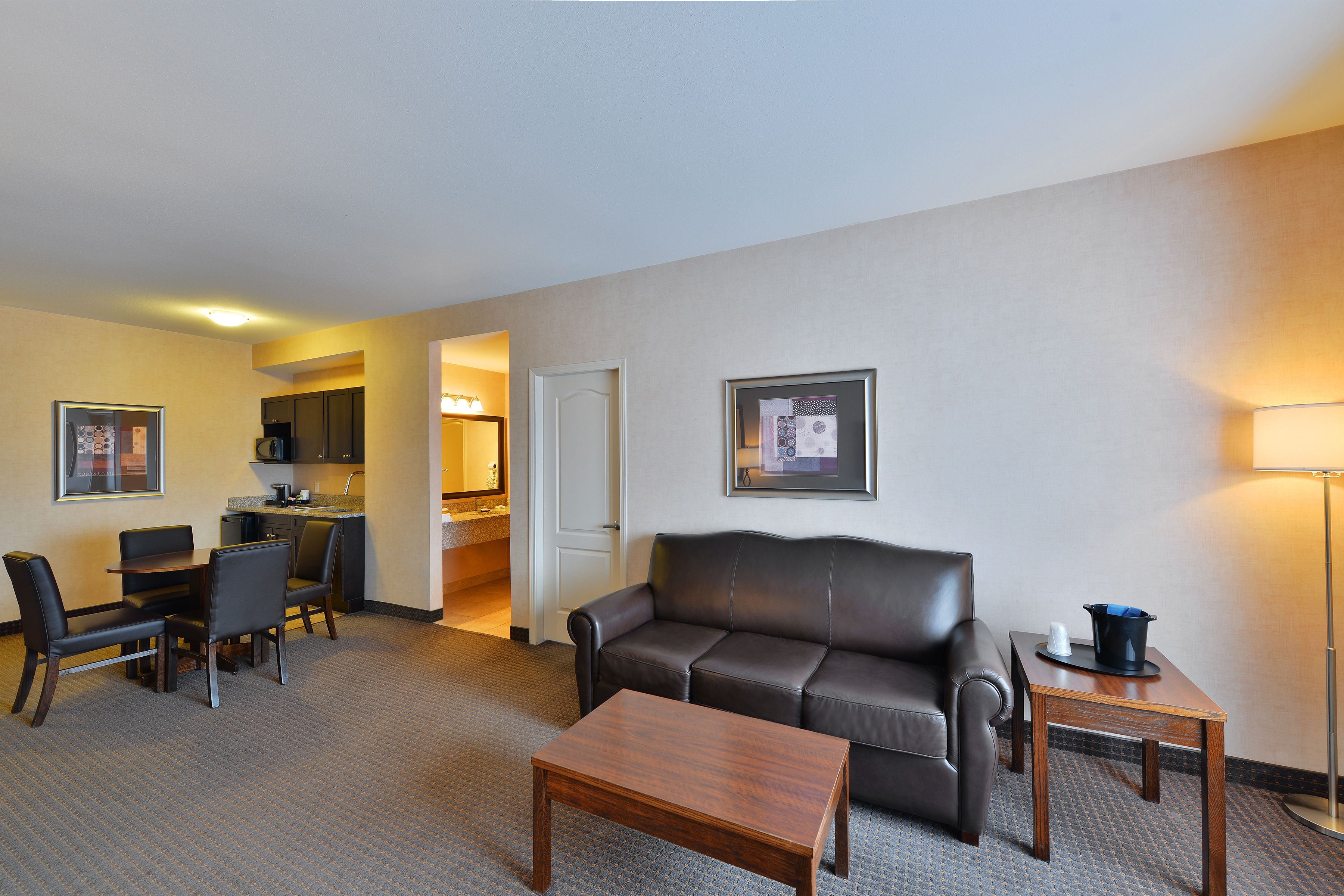 A hotel room with a living area featuring a sofa, a dining table with chairs, a small coffee table, a lamp, and a kitchenette in the background.