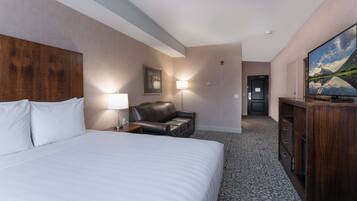 A hotel room with a large bed, a leather chair, a desk with a lamp, a television, and a painting on the wall.