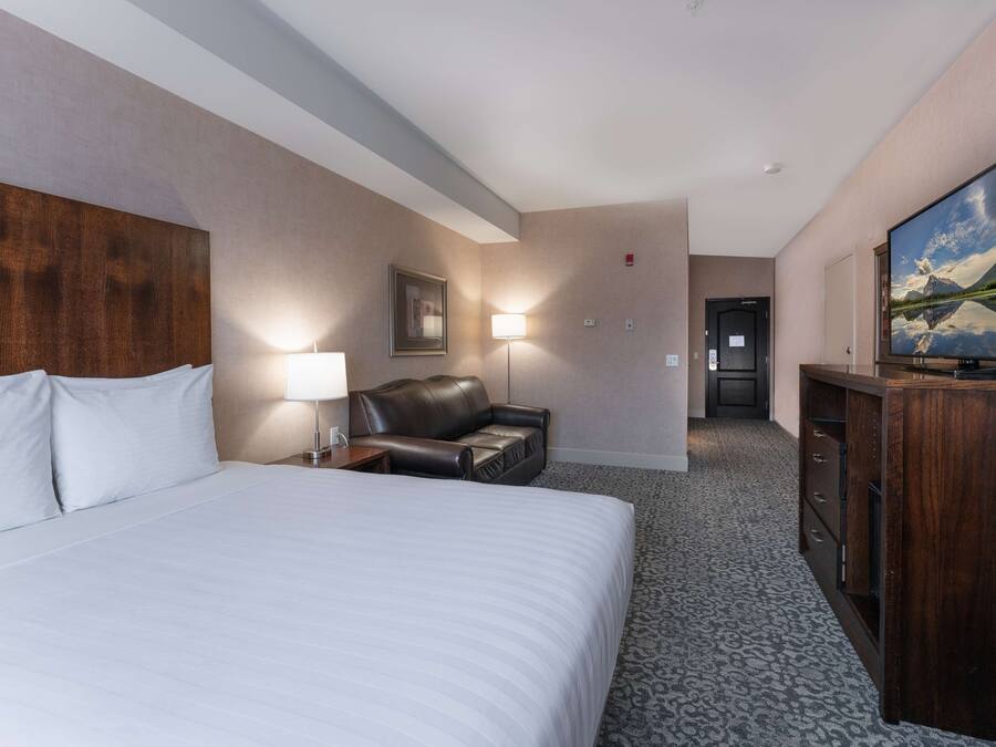 A hotel room with a large bed, a leather chair, a desk with a lamp, a television, and a painting on the wall.