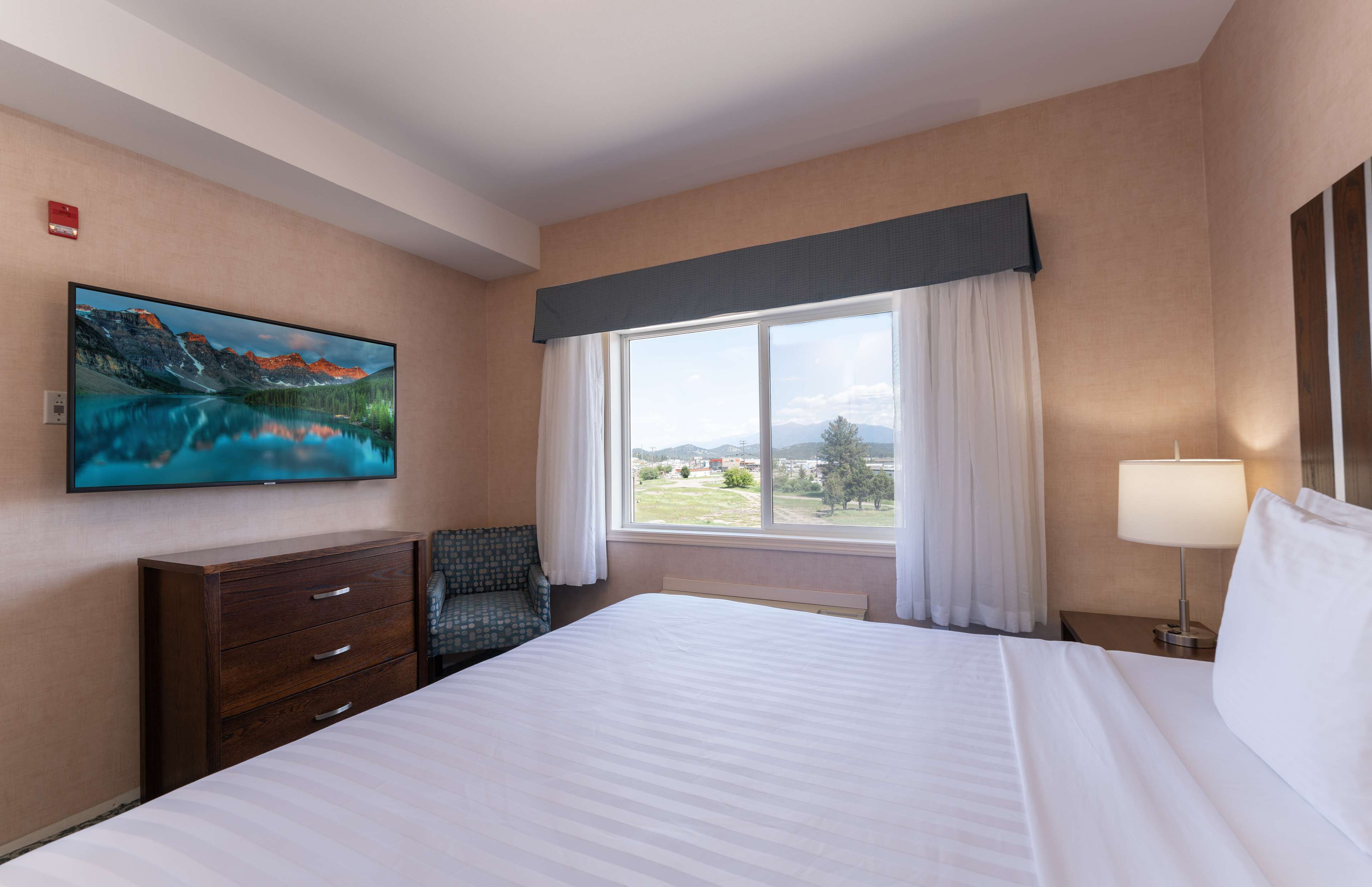 Una habitación de hotel con una cama grande, un aparador, una silla, una ventana con vistas a montañas y un cuadro en la pared.