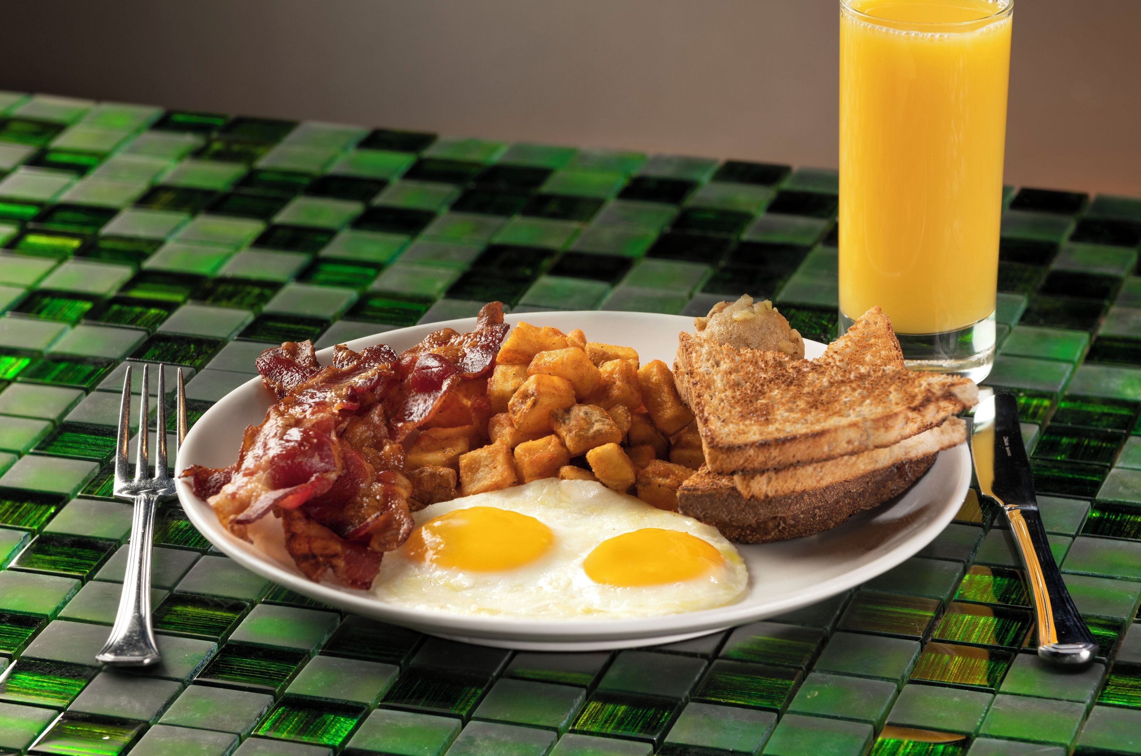 A breakfast plate with eggs, bacon, hash browns, toast, and a glass of orange juice on a checkered table.