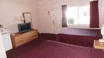 Quarto de hotel com duas camas, uma televisão sobre uma cômoda de madeira, uma porta branca e uma janela com cortinas.
