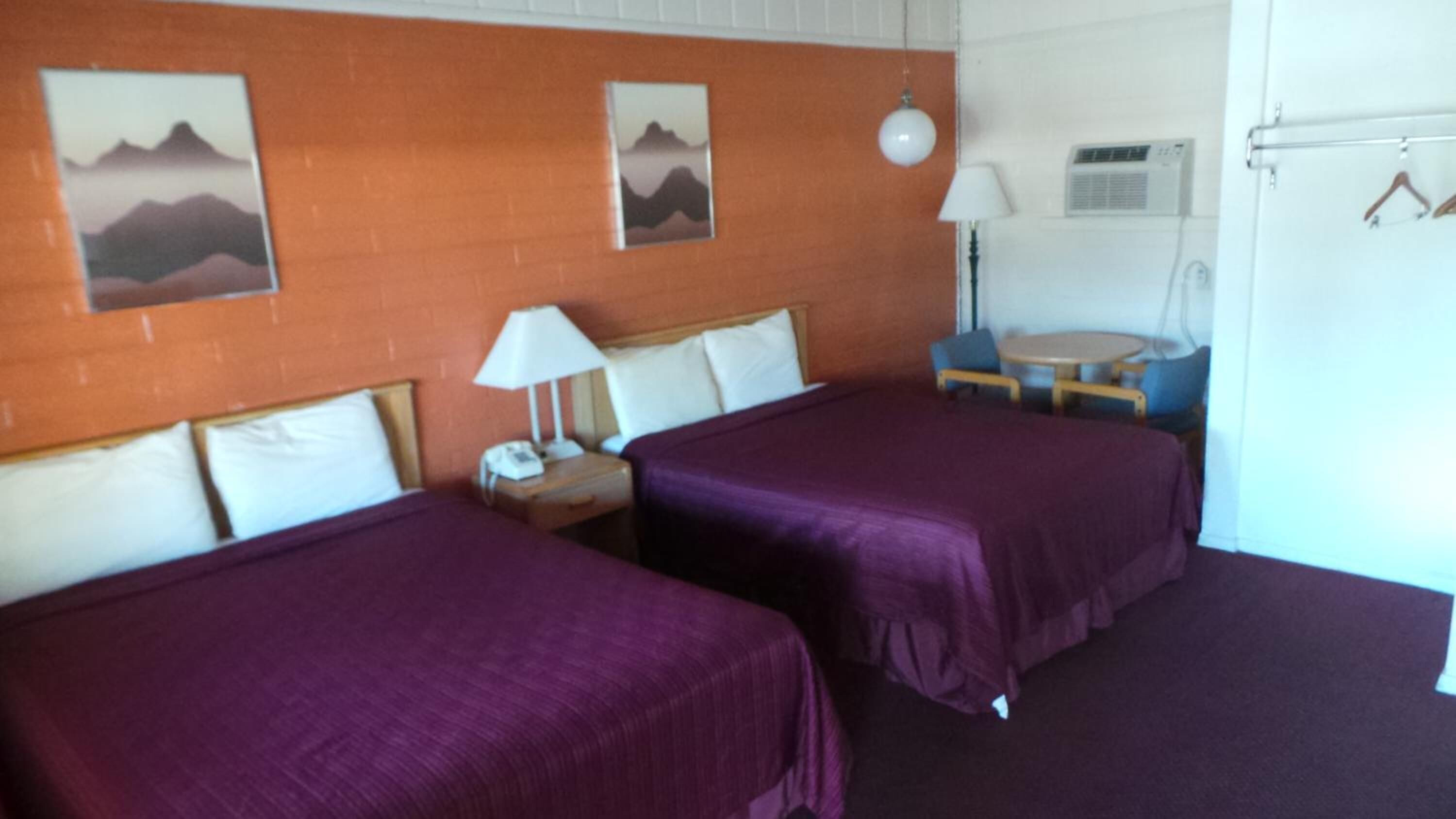 Ett hotellrum med två sängar, ett litet bord, en stol, en lampa och en väggmonterad luftkonditioneringsanläggning.