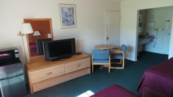 A hotel room with a television on a wooden dresser, a small round table with a chair, a bed with a purple blanket, and a bathroom visible through an open door.