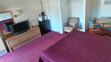 A hotel room with a bed, a television on a wooden dresser, a chair, a small table, and a wall-mounted heater.