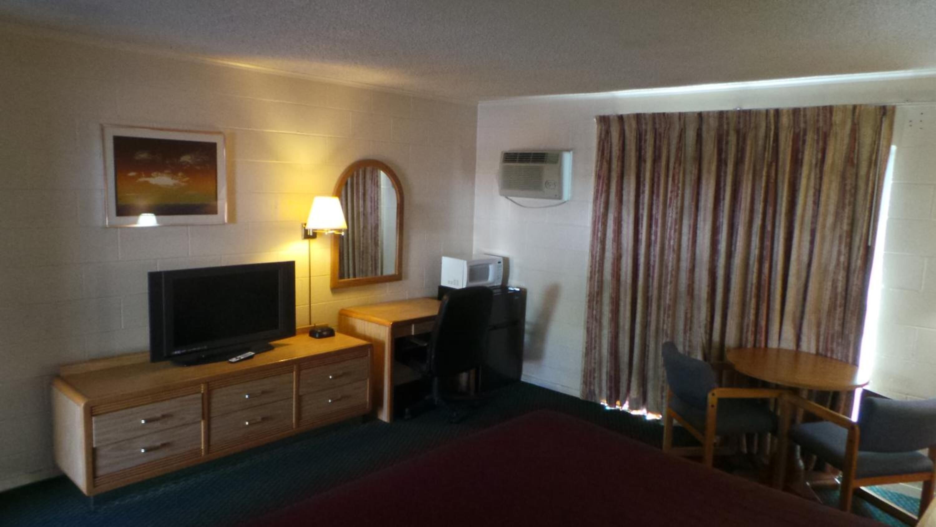 A hotel room with a bed, a TV, a desk with a chair, a lamp, a microwave, and a window with curtains.