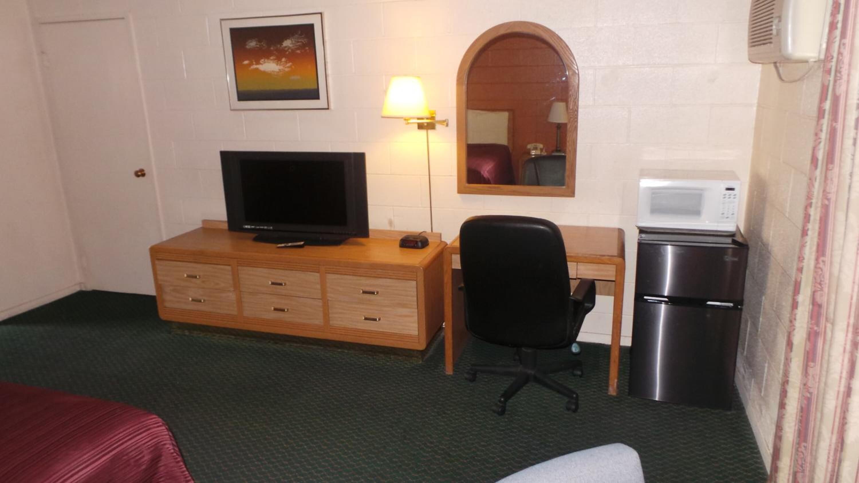 A hotel room with a wooden dresser, a television, a desk with a chair, a microwave, and a refrigerator.