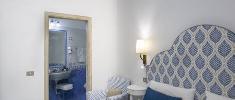 A hotel room with a bed, a chair, a bedside table, a lamp, and a bathroom visible through an open door.