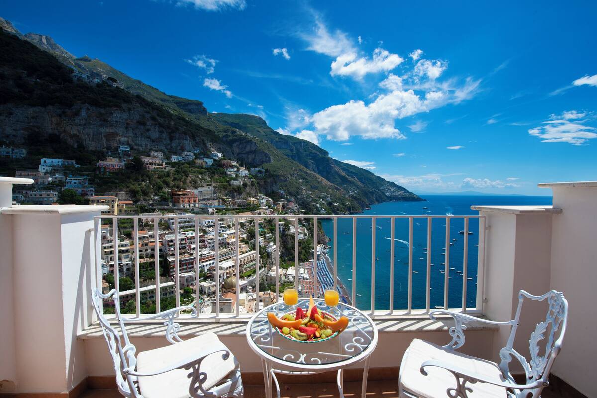 A balcony with a table set for two, overlooking a coastal town and the sea.