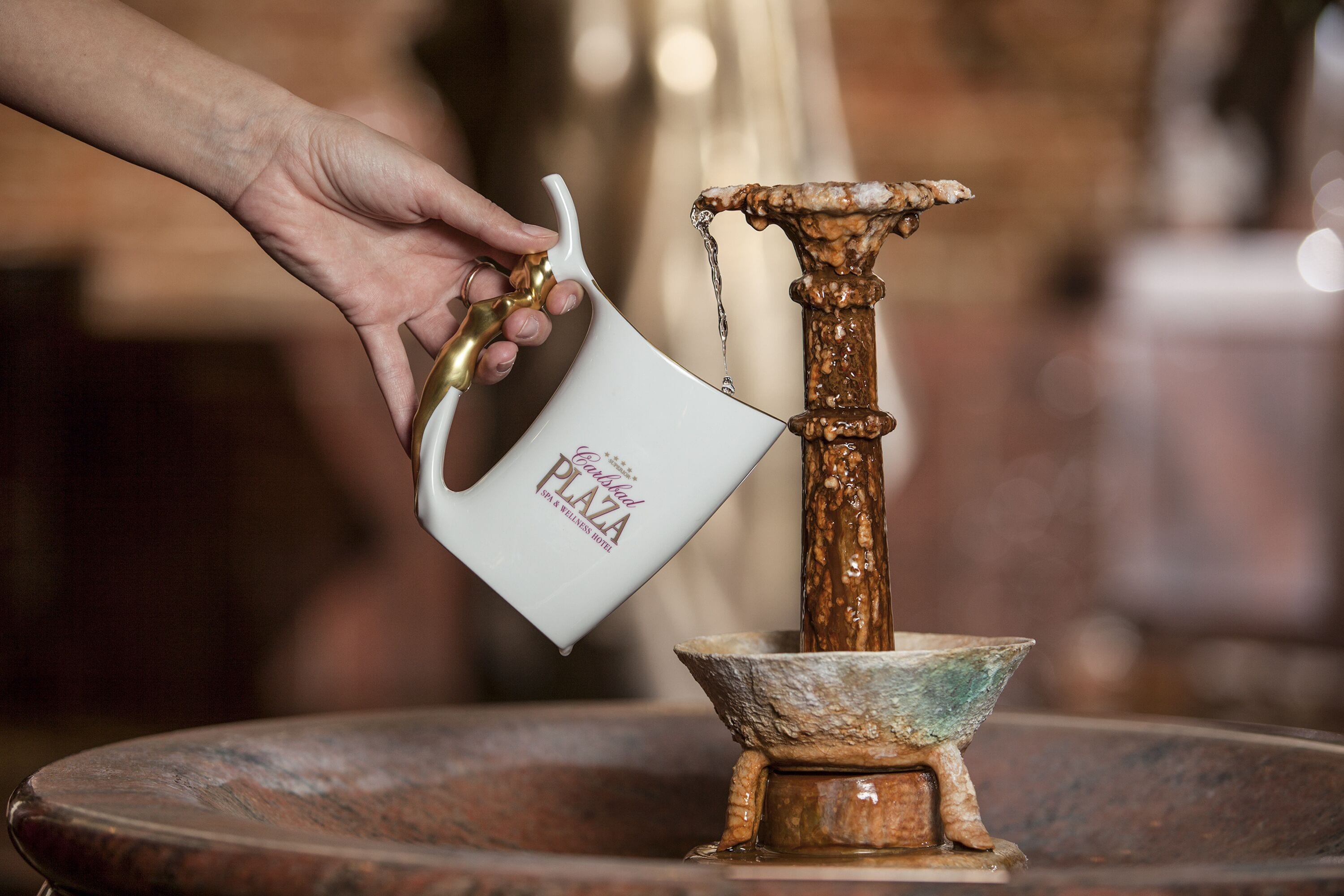 Een hand giet water uit een witte mok in een koperen fontein.