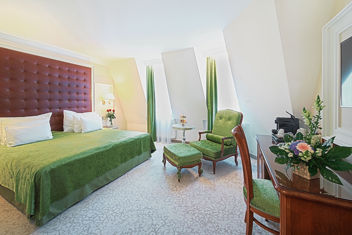 Une chambre d’hôtel avec un grand lit, un fauteuil recouvert d’un tissu vert, une petite table avec une lampe et une plante décorative.
